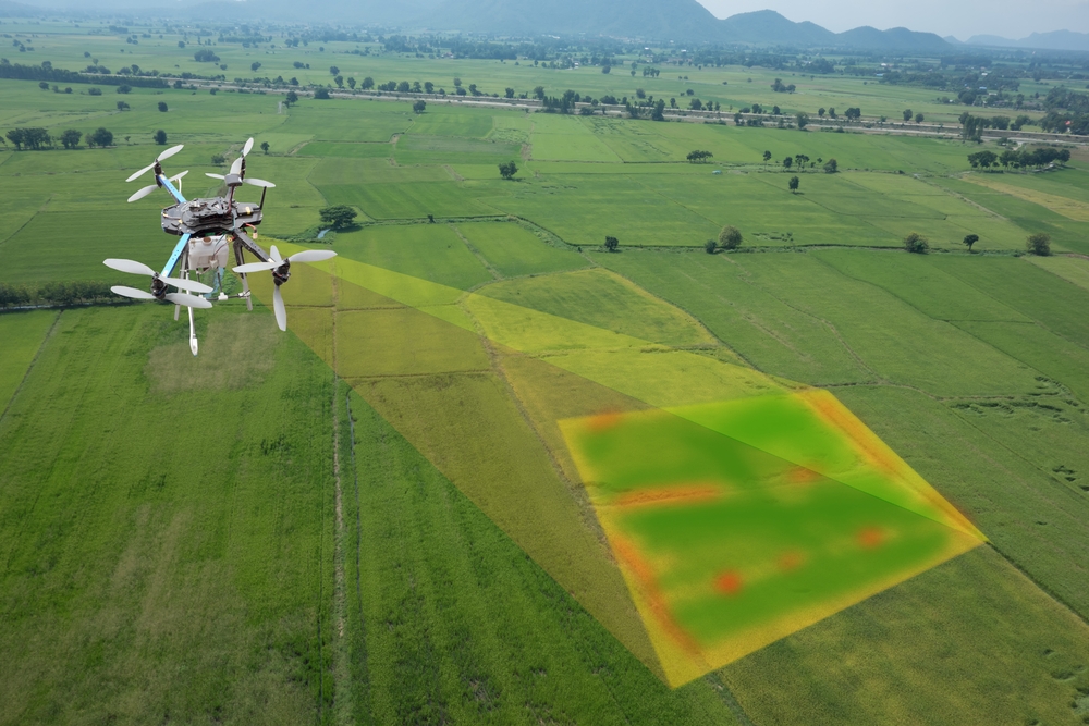 Agricultural inspection aerial drone flies high over flat green fields. A rectangular portion of the field shows in multiple colors, as it would in thermal imaging. The drone is imaging the crop.
