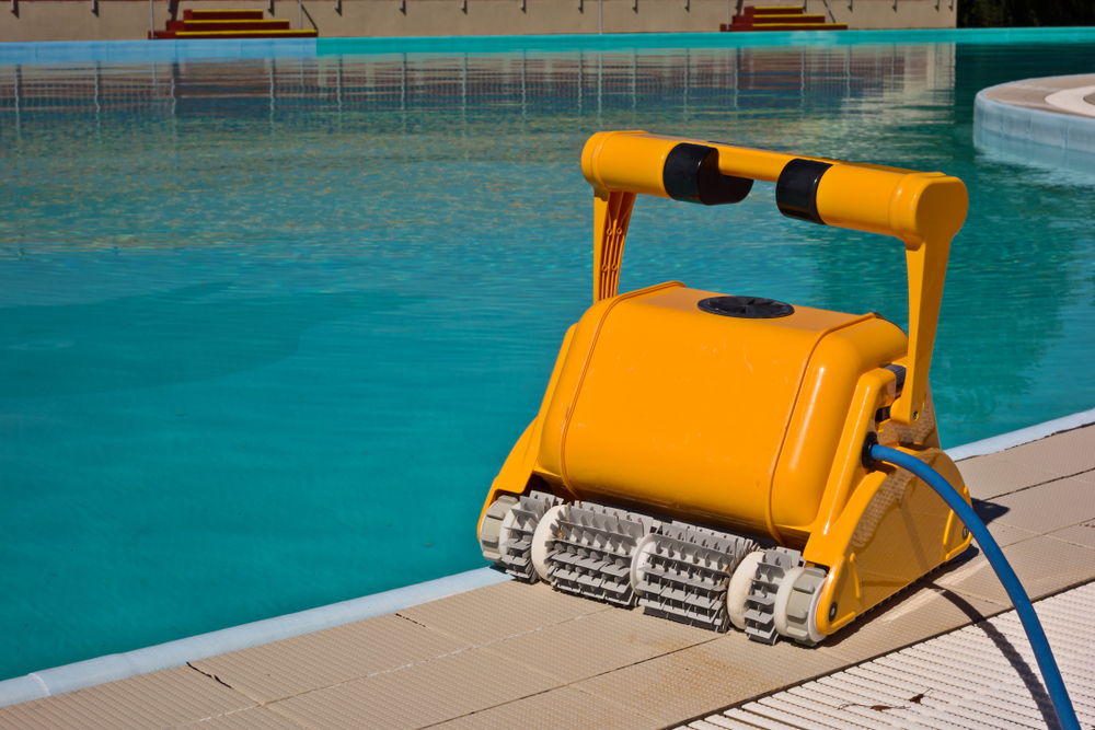A pool cleaning robot rests on concrete next to a pool. A blue hose is attached to the side of the robot. The robot is orange.