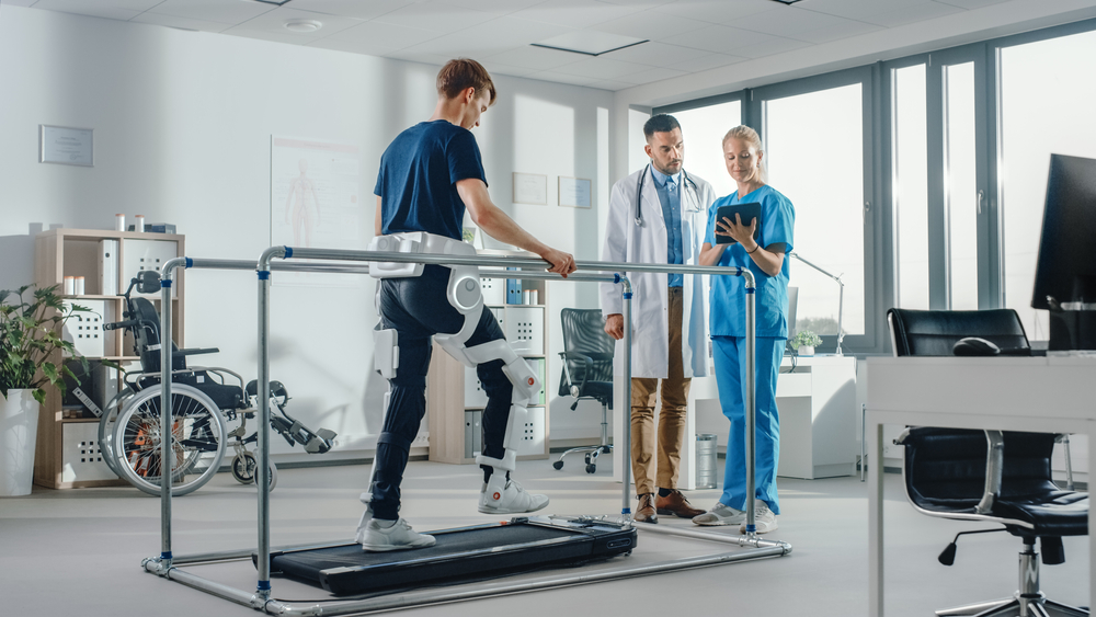 Rehabilitation robotics. A young man stands between handrails on a treadmill. A robotic device is attached to one of his legs. Two healthcare personnel stand nearby and are looking at a tablet.