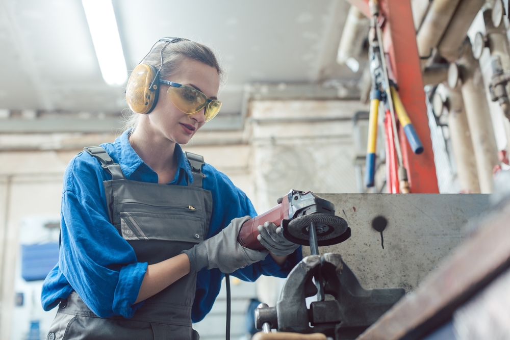 A woman wearing safety glasses, ear protection, and gloves uses a manual angle-grinder to debur a piece of metal held by a vise in a factory environment.
