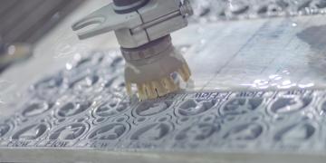 A waterjet cutting robot is shown in action cutting detailed pieces out of a sheet of plastic about 2 inches thick.