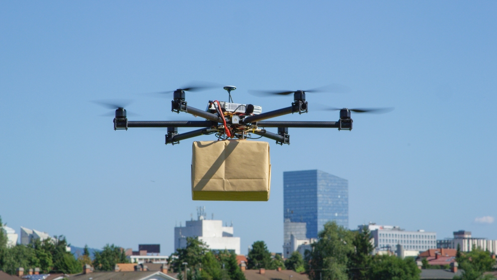 Aerial delivery drone shown hovering, holding a brown box.