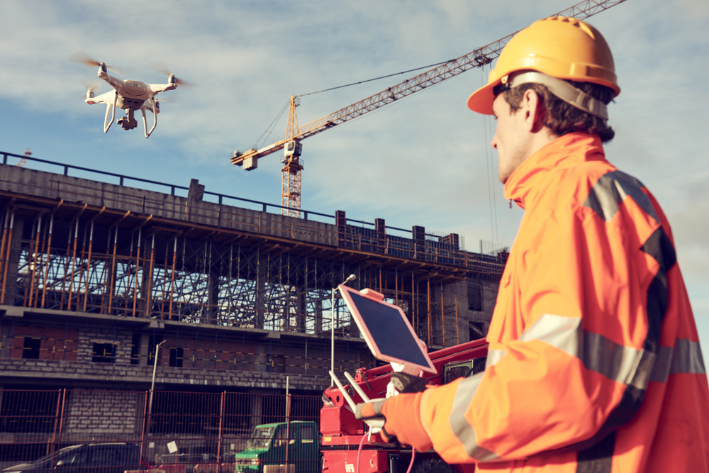 An aerial drone hovers over a building under construction. A man in a hard hard with a drone controller is on the ground looking up at the drone.