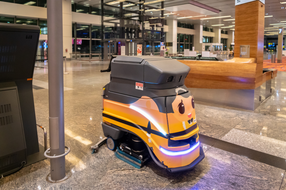 A floor cleaning robot with a painted face on its front. The robot is in an airport or office building, with marble floors and many pillars.
