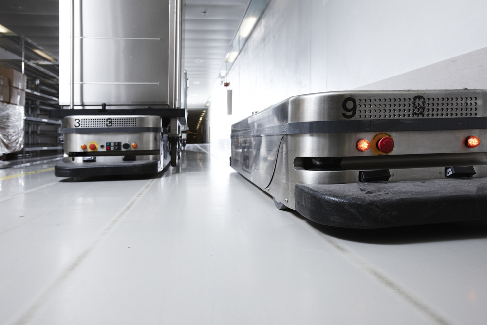 Two autonomous mobile robots in a hospital hallway. Low profile devices with wheels colored silver. One robot has a large metal machine on top of it.