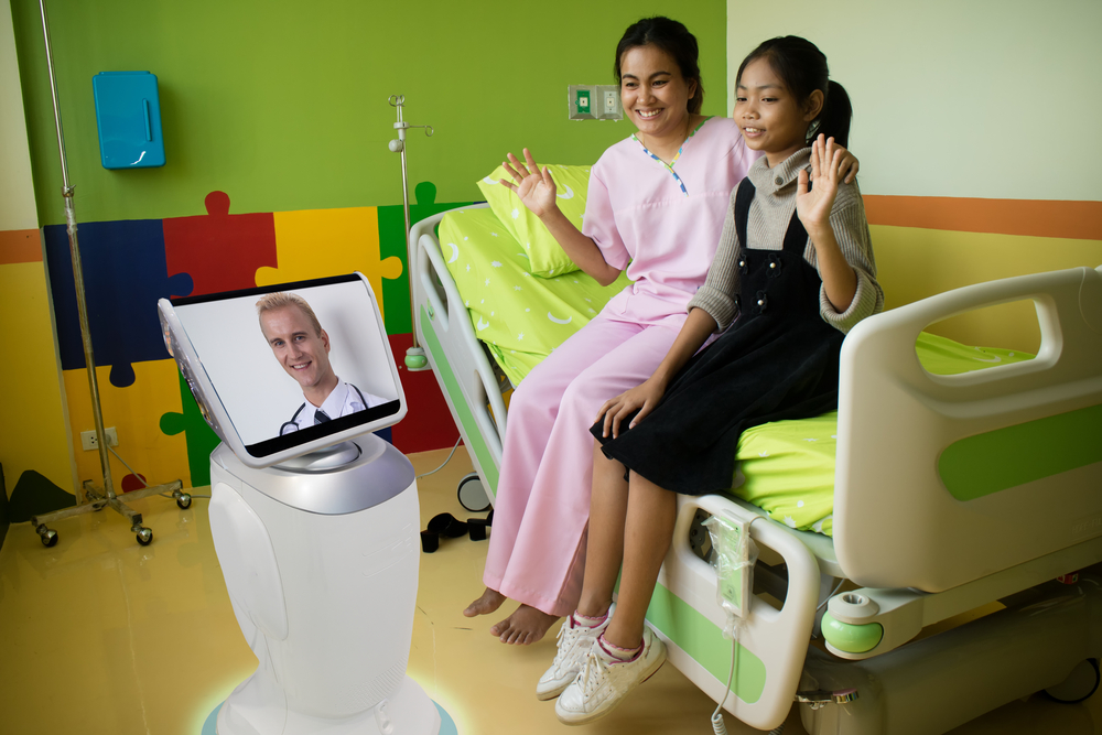 A telepresence healthcare robot is positioned next to a hospital bed with an adult woman and a child sitting on it. Both of the females have their hand up, waving hello to the image of a doctor who appears on the display of the mobile robot. 